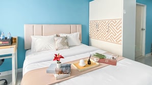 A hotel room with a bed, a bedside table with water bottles, a tray with a book, a potted plant, and a wardrobe. - Neat Studio With Blue Toned Wall At Dramaga Tower Apartment (Dramaga)