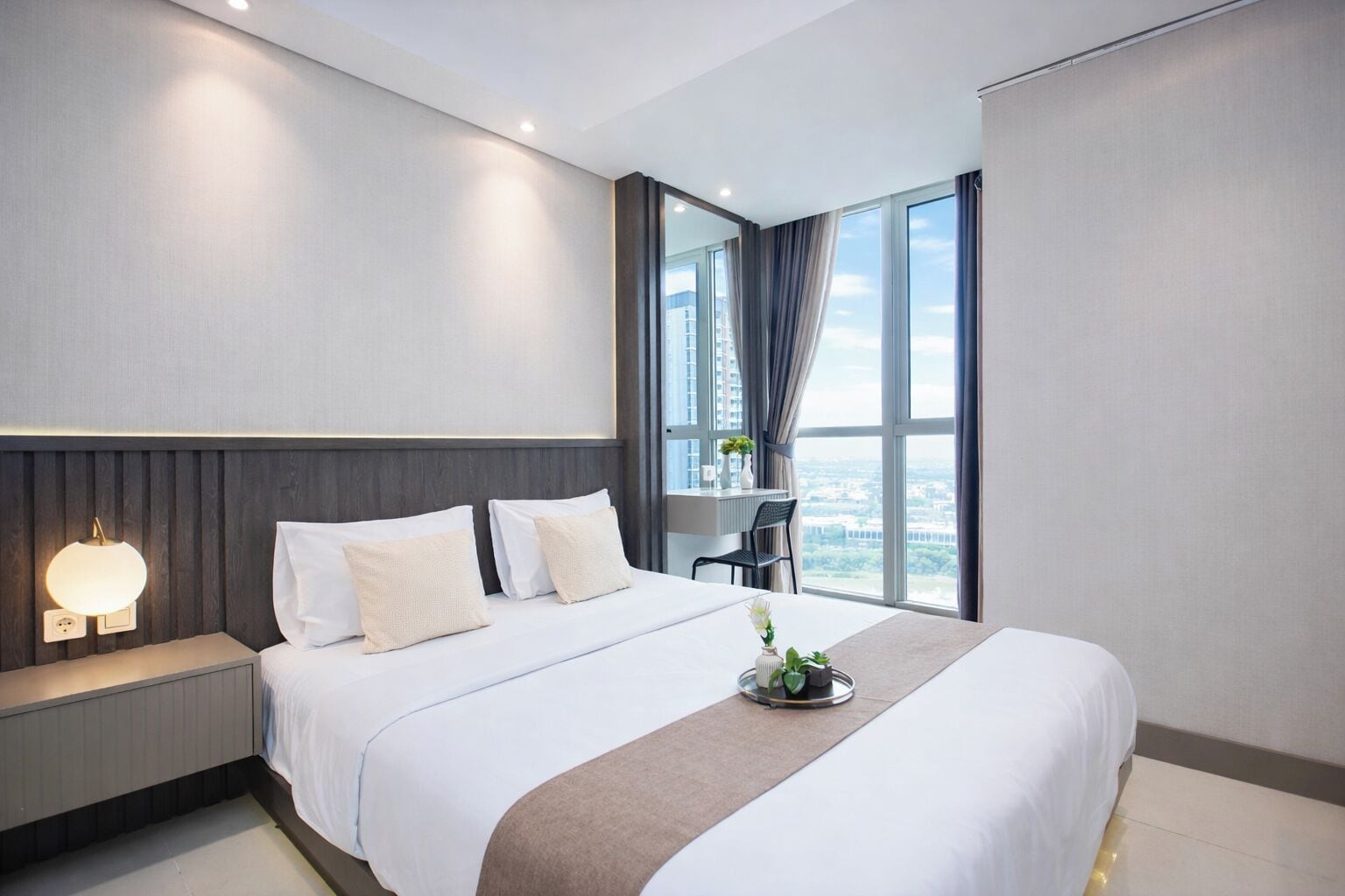 A modern hotel room with a large bed, a bedside table with a lamp, a view of the city through large windows, and a small desk area.
