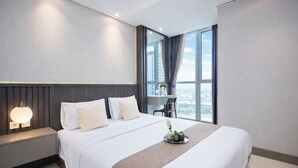 A modern hotel room with a large bed, a bedside table with a lamp, a view of the city through large windows, and a small desk area. - 1Br Homey Areum Residence At Gold Coast (Jakarta)