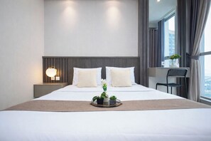 A modern hotel room with a large bed, bedside lamps, a small table with a tray of flowers, and a view of the city through the window. - 1Br Homey Areum Residence At Gold Coast (Jakarta)