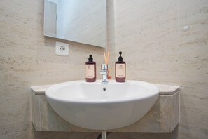 A bathroom with a white sink, two bottles of body wash, a mirror, and a wall socket. - 1Br Homey Areum Residence At Gold Coast (Jakarta)