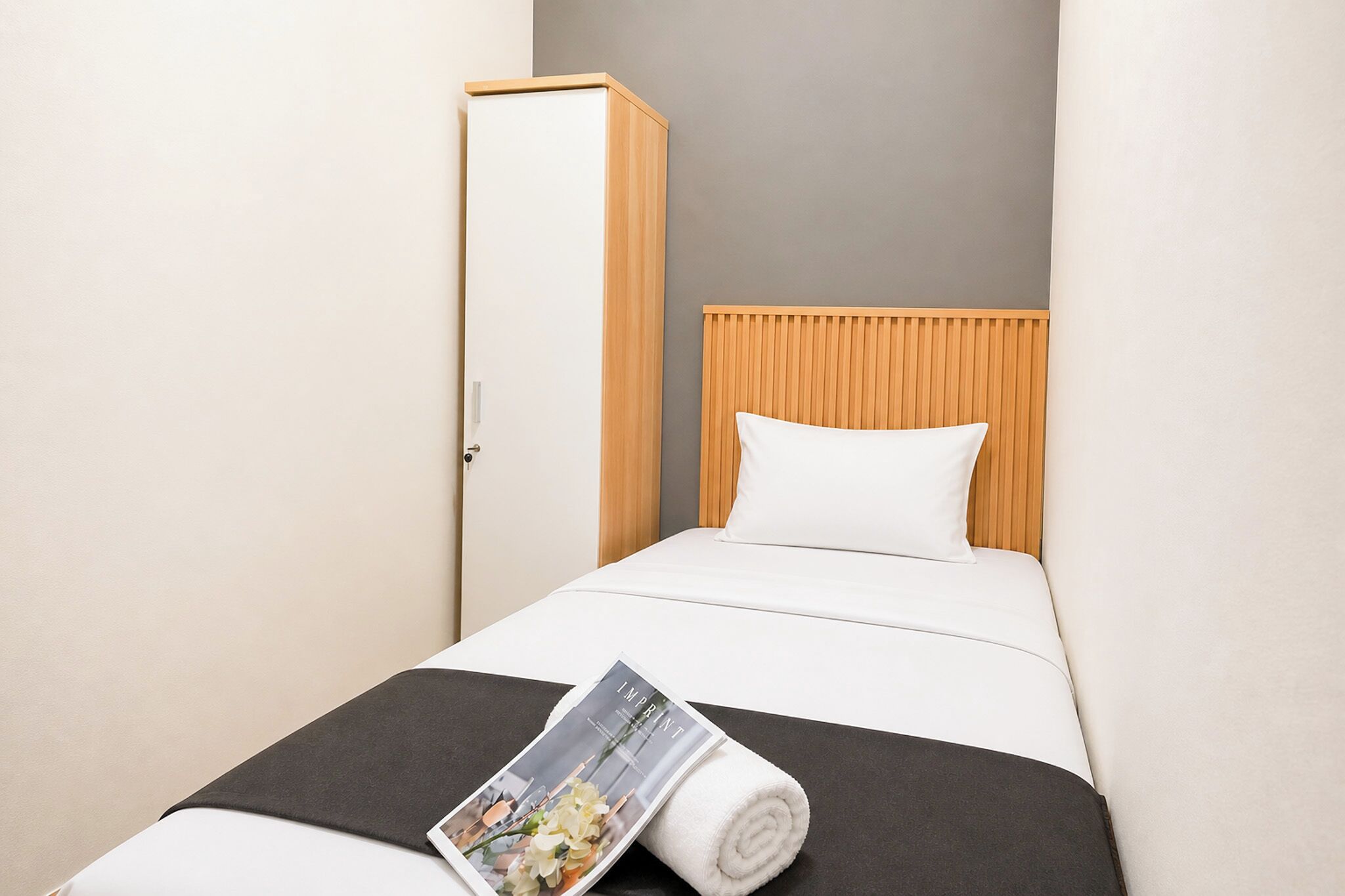 A hotel room with a bed, a wooden headboard, a white pillow, a black bedspread, and a rolled-up magazine on the bed.