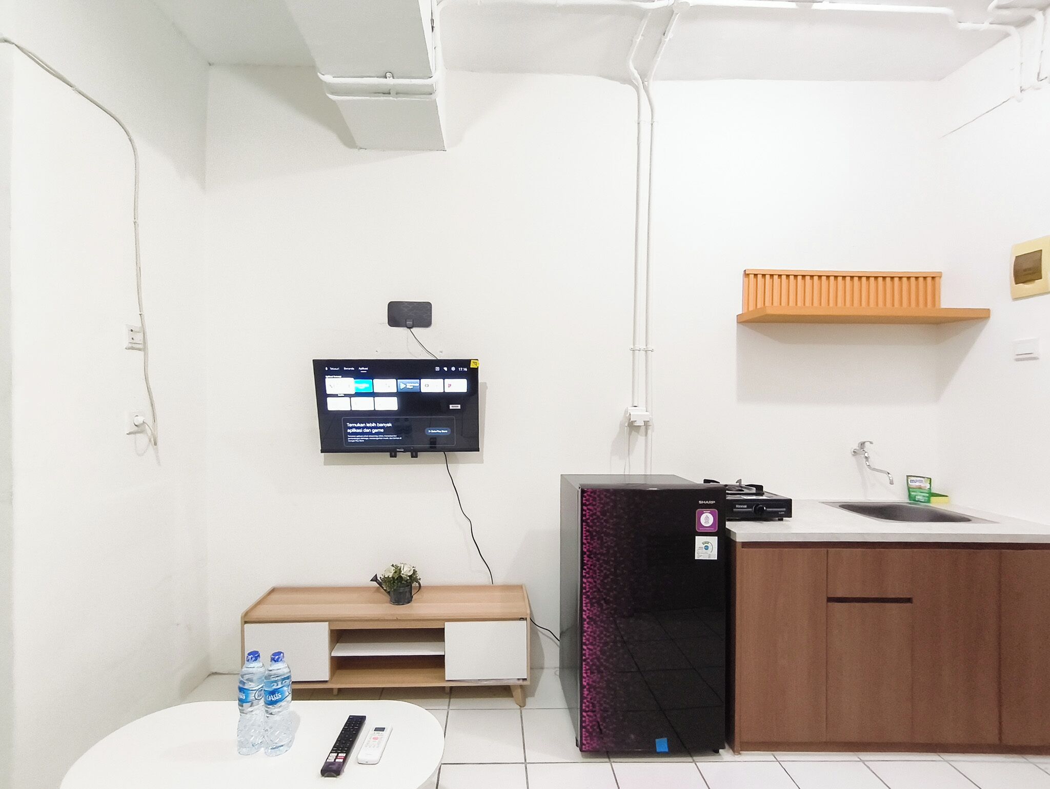 A modern room with a flat-screen TV mounted on the wall, a small wooden cabinet, a white round table with a remote control, and a water bottle.