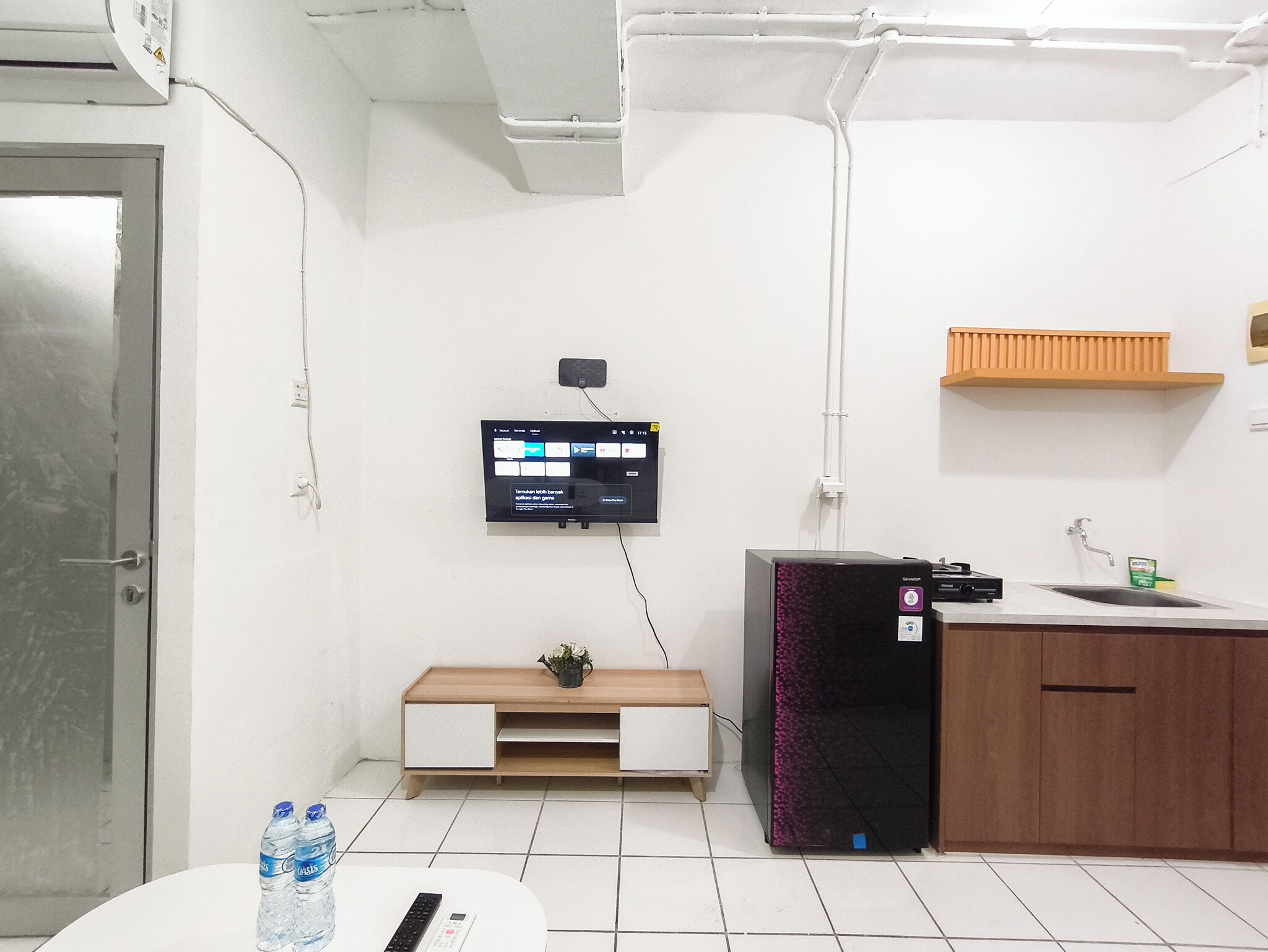 A modern office space with a flat-screen TV mounted on the wall, a wooden bench, a refrigerator, a sink, and a door.