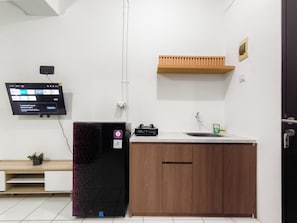 A kitchen with a refrigerator, a sink, a wooden cabinet, a wall-mounted TV, and a small plant. - Practical 2Br For 3 Pax Near Braga At Grand Asia Afrika Apartment (Bandung)