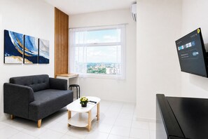 A modern room with a grey sofa, a small wooden table with a plant, a large window with a city view, and a mounted TV displaying a welcome message. - Practical 2Br For 3 Pax Near Braga At Grand Asia Afrika Apartment (Bandung)