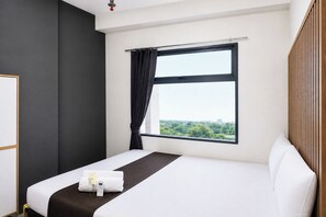 Ein Hotelzimmer mit großem Fenster, einem Bett, einem Nachttisch mit Fernbedienung und Blick auf Grünflächen.