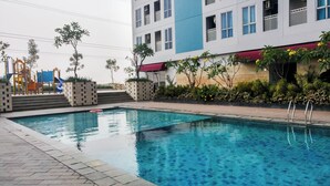 A swimming pool with a clear blue water surface, surrounded by a paved walkway. There are steps leading up to a building with a blue and white facade, and a playground with colorful equipment is visible in the background. - Minimalist And Restful Studio Room At Apartment Grand Dhika City (Bekasi)