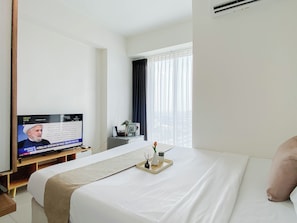 A hotel room with a bed, a TV on a stand, a window with curtains, and a bedside table with a vase and a tray. - Practical Studio For 2 Pax At Grand Kamala Lagoon Apartment (Bekasi)