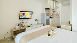 A hotel room with a bed, a TV mounted on the wall, a kitchenette, and a small bedside table with a vase and a tray. - Practical And Restful Studio Room At Apartment Grand Kamala Lagoon (Bekasi)