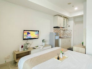 A hotel room with a bed, a TV mounted on the wall, a kitchenette, and a small bedside table with a vase and a tray. - Practical And Restful Studio Room At Apartment Grand Kamala Lagoon (Bekasi)