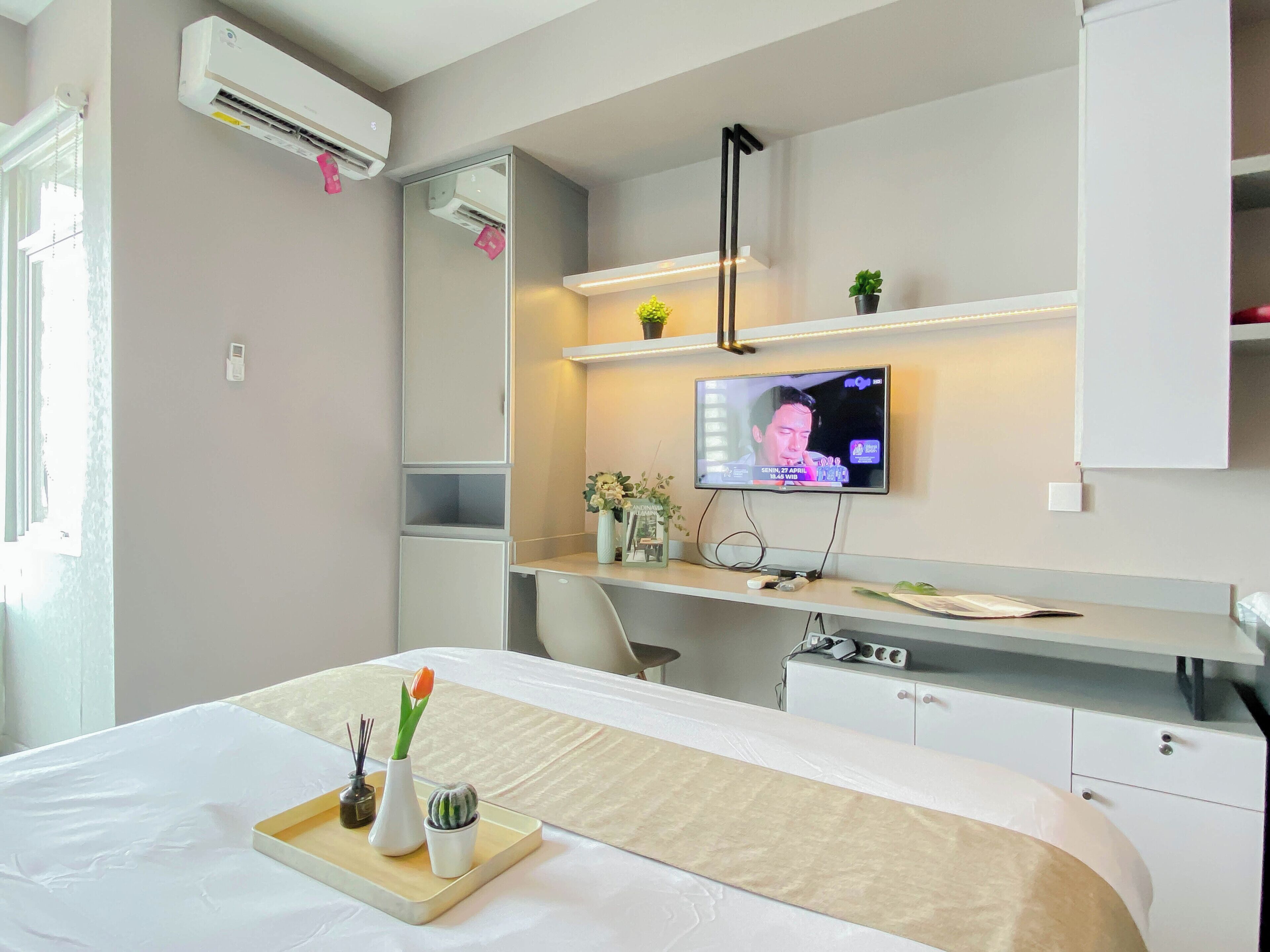 A hotel room with a bed, a desk with a TV, air conditioning, and a small plant.