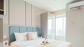 A modern hotel room with a large bed, a bedside table, a nightstand, a window with a city view, and an air conditioning unit. - Restful Stay In Studio Room At Apartment Grand Kamala Lagoon (Bekasi)