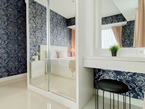 A bedroom with a glass shower, a bed with white bedding, a bedside table with a lamp, and a small potted plant. - Classic 1Br With Extra Room At Apartment Grand Kamala Lagoon (Bekasi)