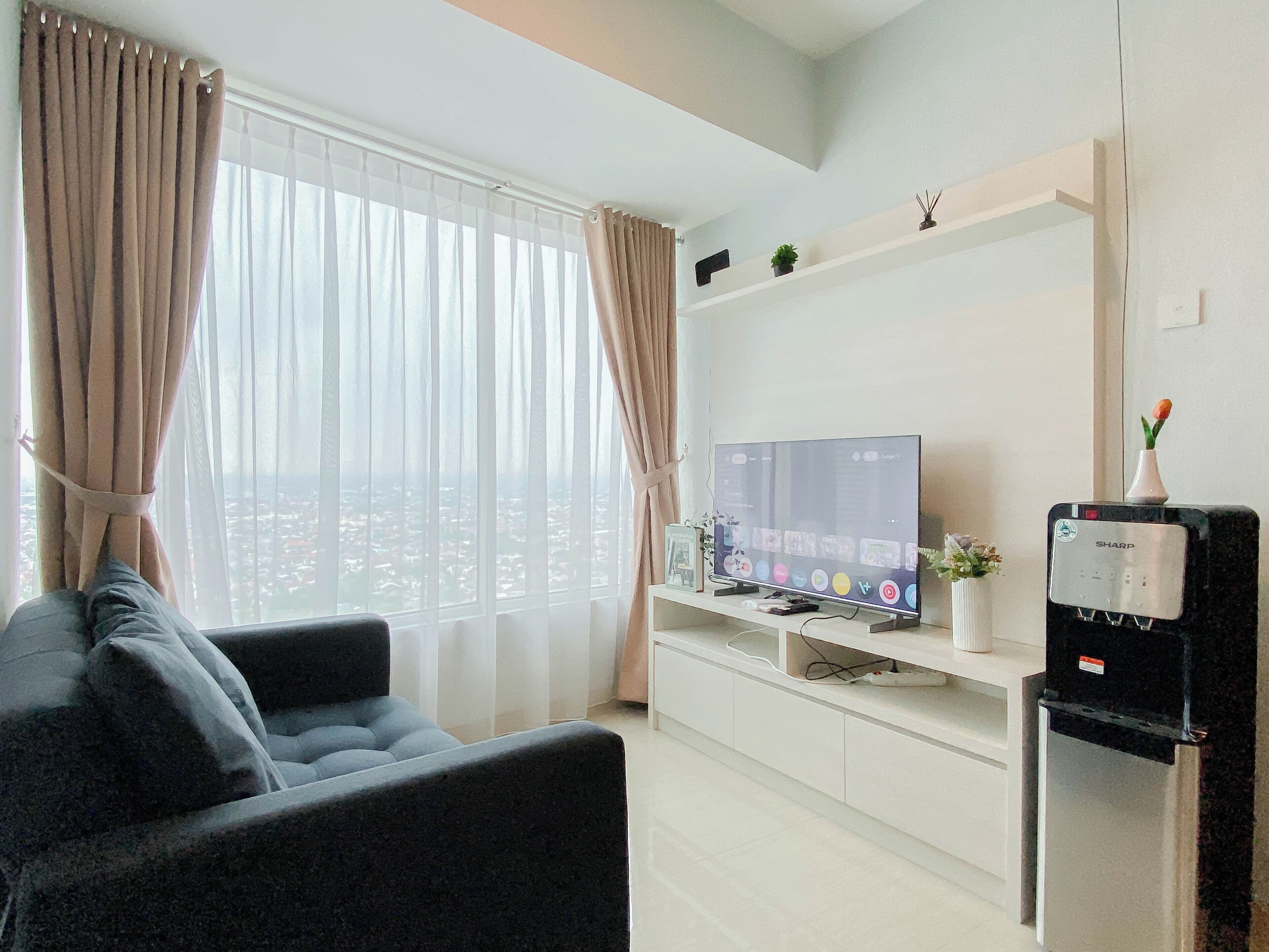 Un salon moderne avec un canapé, un téléviseur à écran plat, un distributeur d’eau et une vue sur la ville à travers des rideaux transparents.