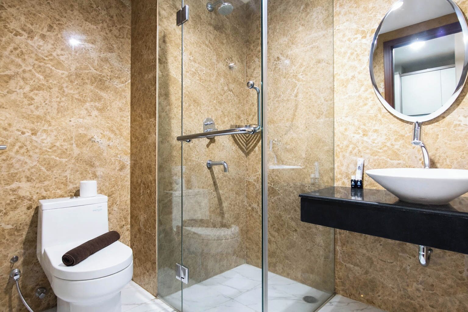 A modern bathroom with a glass-enclosed shower, a white toilet, a black sink with a round mirror, and marble walls.