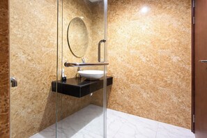 A bathroom with a glass shower enclosure, a marble wall, a black countertop, a white sink, and a round mirror. - 2 Pax Versatile Studio At Grand Sungkono Lagoon Apartment (Surabaya)