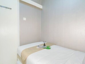 A single bed with a white mattress and a beige blanket, a small table with a green object, and a white wall with a light switch. - Lovely Urban Living In 2Br At Apartment Green Pramuka City (Jakarta)