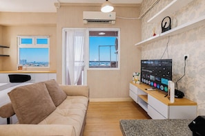 A modern living room with a sofa, a TV, a desk, and a window with a city view. - Classic And Restful Studio Room At Apartment Green Pramuka City (Jakarta)