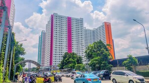A multi-story building with a prominent pink vertical stripe, surrounded by parked cars and greenery. - Classic And Restful Studio Room At Apartment Green Pramuka City (Jakarta)