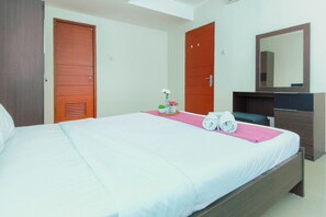 A hotel room with a bed, a wooden wardrobe, a mirror, and a small table with a vase and flowers. - Cozy Living 2Br Apartment At High Point Serviced (Surabaya)