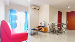 Una sala de estar con un sofá rojo, un televisor sobre un mueble de madera, una mesa de comedor con sillas y un ventanal con cortinas azules.