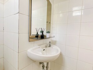 Een badkamer met witte tegels, een wit wastafelmeubel, een spiegel en een plankje met een cactus en een rietverspreider.