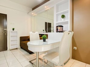 A modern dining area with a glass table, white chairs, a brown sofa, a white shelving unit, and a compact kitchen area with a rice cooker. - Comfortable And Decent 2Br At Apartment Kemang View (Bekasi)