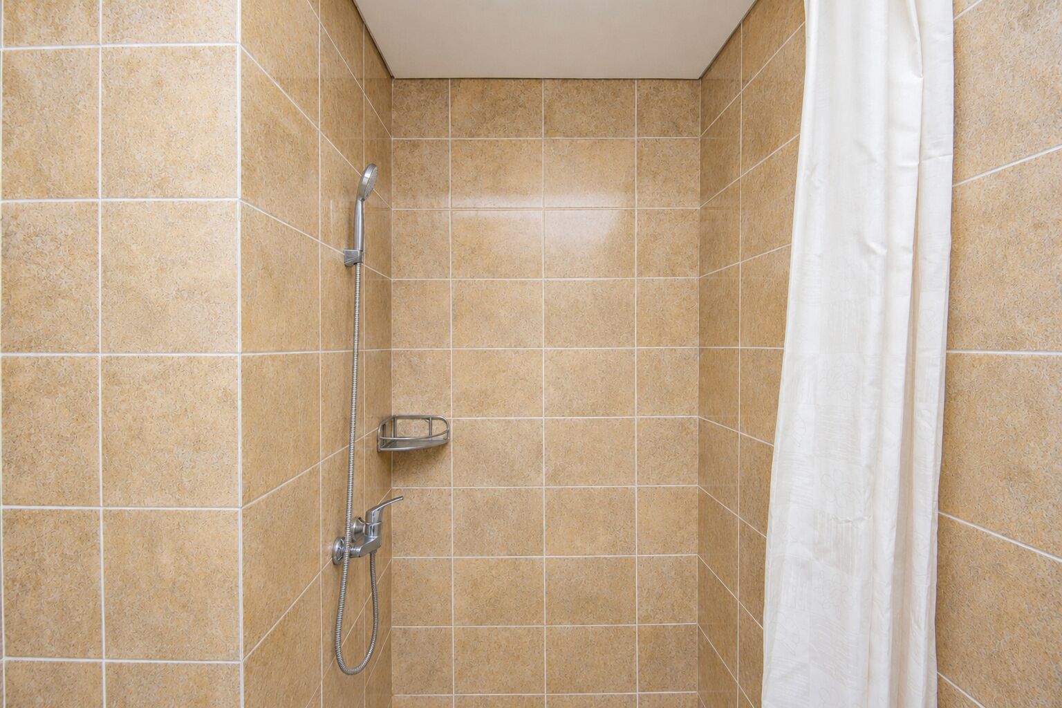 A shower area with beige tiles, a showerhead, a handheld showerhead, and a white shower curtain.