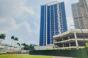 A high-rise building with a blue facade, a parking structure, and a green area with palm trees. - Serene Loft Studio At Kingland Avenue Apartment (South Tangerang)