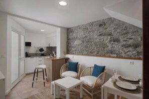 A modern kitchen with white cabinets, a stone accent wall, and a dining area with wicker chairs and a white table. - Sun Hill – Comfort in the South of the Island (Ponta do Sol)