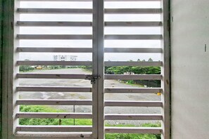 A view through a window with horizontal blinds, a building in the background, and a green area with trees. - Comfy And Nice Studio (No Kitchen) At Apartment Kubikahomy (Pagedangan)