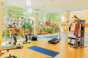 Ein Fitnessraum mit Trainingsgeräten, einer blauen Matte und Blick auf ein Schwimmbecken und Grünflächen.