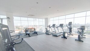 Academia com equipamentos de ginástica, grandes janelas e vista para a cidade.
