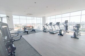 A gym with exercise equipment, large windows, and a city view. - Comfy Studio At Kyo Society Apartment (Surabaya)