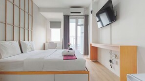 A hotel room with a bed, a TV mounted on the wall, an air conditioning unit, a desk with a chair, and a window with curtains. - Zen Style Studio At Kyo Society Apartment (Surabaya)