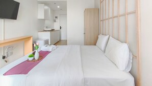 A hotel room with a bed, a bedside table with a vase of flowers, a small desk with a chair, a wardrobe, and a TV mounted on the wall. - Zen Style Studio At Kyo Society Apartment (Surabaya)