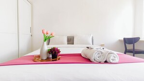 Een netjes opgemaakt bed met witte lakens, een roze sprei, een witte vaas met roze bloemen en een kleine potplant op een nachtkastje.