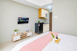 A modern hotel room with a bed, a TV mounted on the wall, a small kitchenette, and a bedside table with a plant. - Restful Studio With City View At Royal Sentul Park Apartment (Bogor)