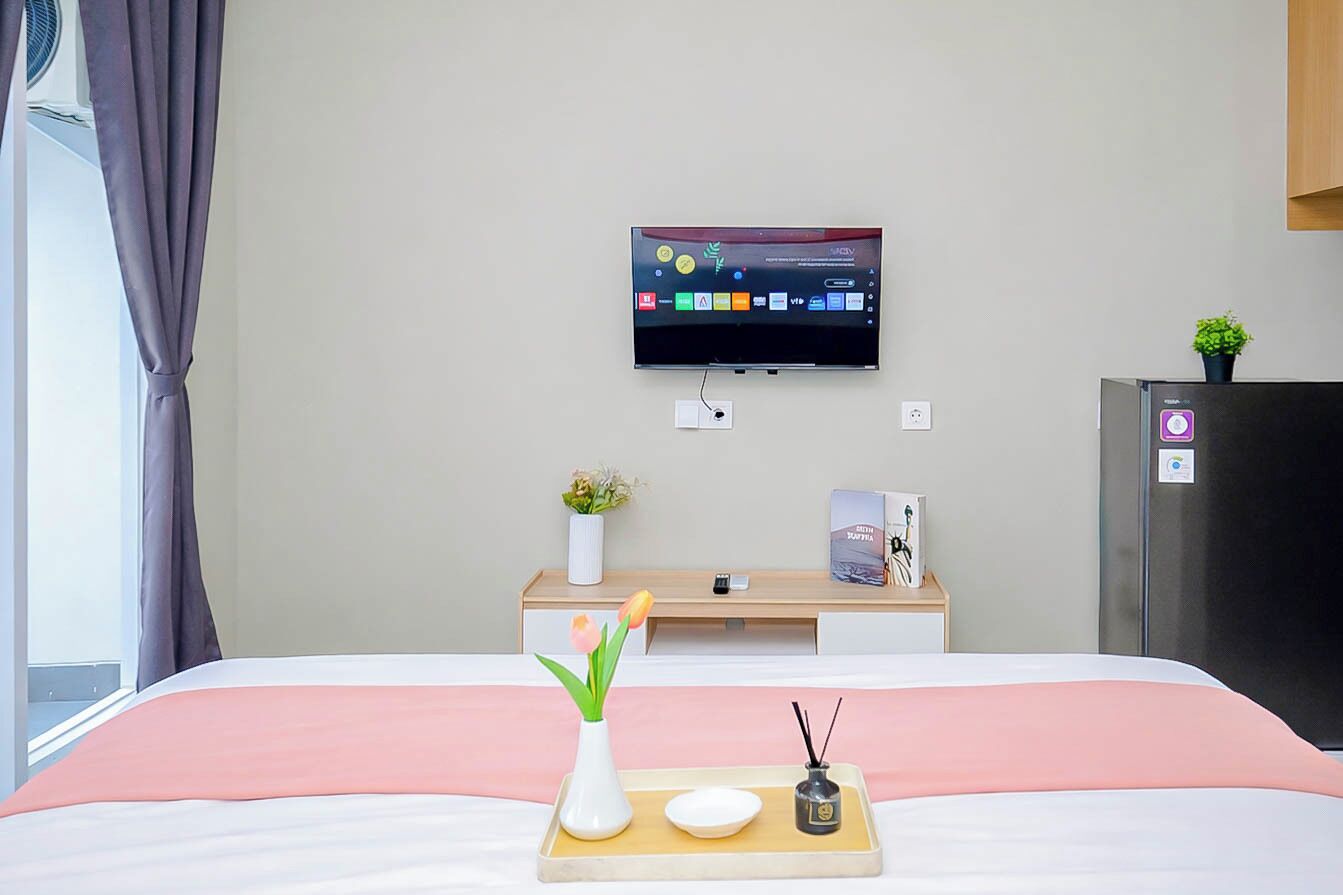 Habitación de hotel con cama, televisión montada en la pared, una pequeña mesa de escritorio con planta y refrigerador.