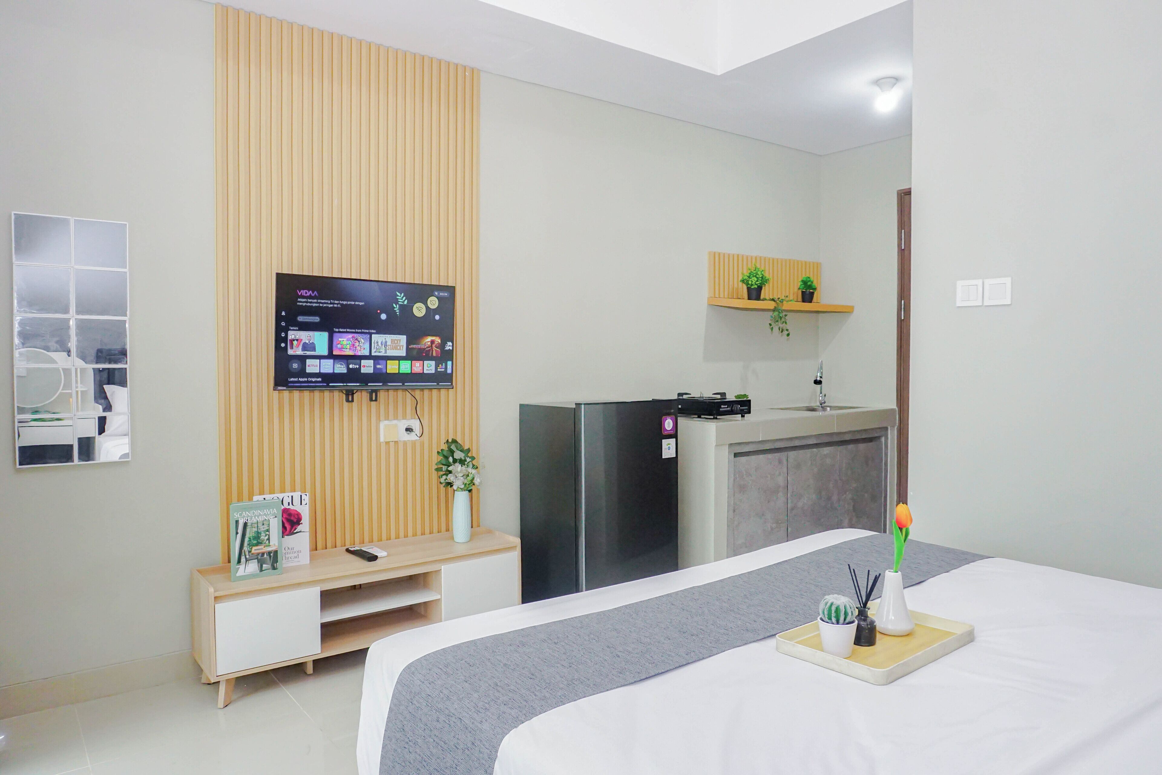 A hotel room with a bed, a TV mounted on a wooden panel, a small kitchenette, and a shelf with plants.