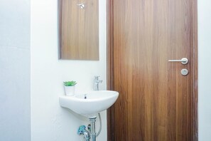 A bathroom with a wooden door, a white sink, a small potted plant, and a mirror above the sink. - Serene Studio At Royal Sentul Park Apartment (Bogor)