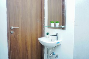 A bathroom with a wooden door, a white sink with a mirror above it, and two potted plants on a shelf. - Comfortable Living Studio At Apartment Lrt City Sentul - Royal Sentul Park (Bogor)