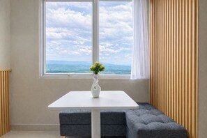 Ein Zimmer mit weißem Tisch, einer Blumenvase, einer Bank und einem Fenster mit Blick auf den Himmel und die Berge.