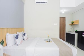 A hotel room with a bed, bedside table, a small desk with a chair, a mirror, a plant, and a door. - Neat And Restful Studio At Apartment Lrt City Sentul - Royal Sentul Park (Bogor)