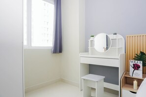 A bedroom with a white desk, a small white stool, a bedside table with a book, and a window with a curtain.