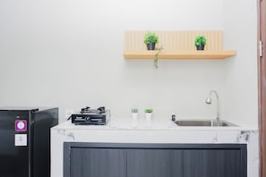 Uma cozinha com bancada branca, geladeira preta, prateleira de madeira com plantas em vasos, pia e torneira.