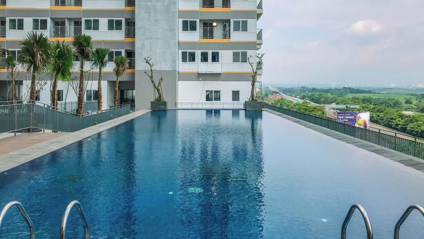 A modern building with a swimming pool, surrounded by palm trees and a view of distant greenery. - Neat Studio For 2 Pax At Royal Sentul Park Apartment (Bogor)