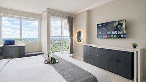 A hotel room with a bed, a desk with a laptop, a TV mounted on the wall, and a balcony with a view. - Neat Studio For 2 Pax At Royal Sentul Park Apartment (Bogor)