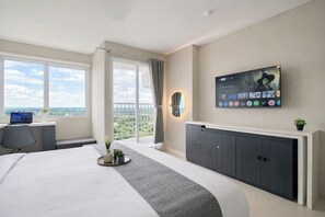 A hotel room with a bed, a desk with a laptop, a TV mounted on the wall, and a balcony with a view. - Neat Studio For 2 Pax At Royal Sentul Park Apartment (Bogor)
